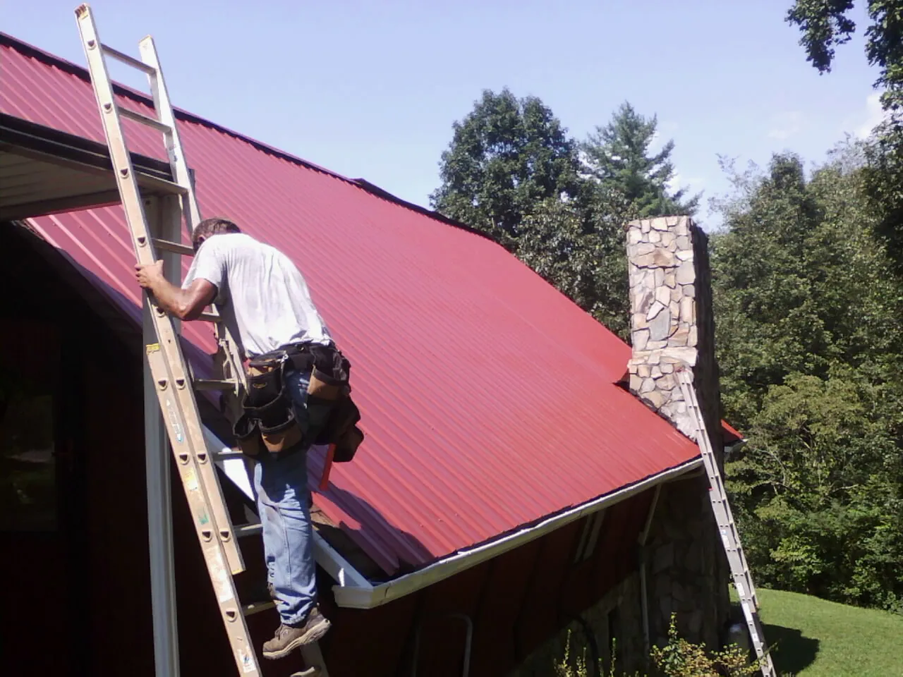 Roofer on a ladder beside a red metal roof and stone chimney — Soffit & Fascia Repair by Roofing Brothers in Dillsboro